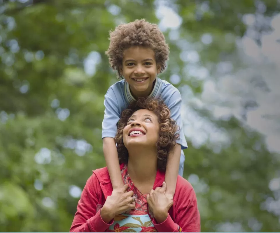 Mulher negra com criança apoiada sobre seus ombros, ambos sorridentes